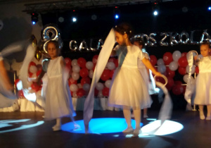 Na scenie tańczą dziewczynki w białych sukienkach z szarfami.